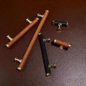May include: An assortment of cabinet pulls and handles with woven leather and brass accents. The collection includes long bar pulls and shorter T-shaped pulls in shades of brown and black. The hardware is set against a brown background.