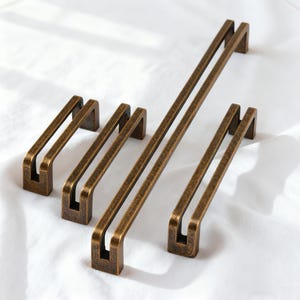 May include: A collection of bronze-colored cabinet pulls in various sizes. The pulls have a rectangular design with a double bar handle. The hardware is arranged on a white surface, showcasing their antique finish.
