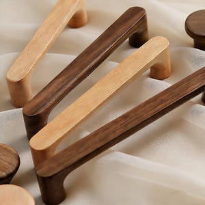 May include: A selection of wooden cabinet handles in different shades of brown and natural wood. The handles are rectangular with curved supports, displaying the wood grain. Some are light tan, others a dark brown. The handles are likely to be used for furniture.