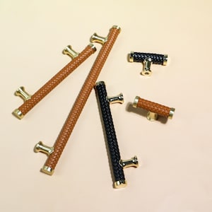 May include: A collection of cabinet hardware featuring woven leather handles in brown and black, with gold-toned accents. The hardware includes long bar pulls and shorter T-shaped knobs, suitable for drawers and cabinets.