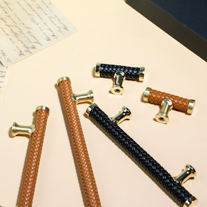 Op de afbeelding: Een assortiment kastgrepen met een geweven lederen design. De grepen zijn in bruin- en zwarttinten, met goudkleurige hardware. De kastgrepen zijn in verschillende maten en vormen, geschikt voor meubels of kasten.
