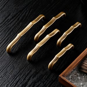 May include: Six antique brass cabinet pulls with a bamboo-inspired design. The pulls have a curved handle and a warm, golden hue. They are arranged on a dark, textured surface, suggesting a display of hardware for furniture or cabinetry.