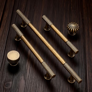 Può includere: Un assortimento di ferramenta per mobili in ottone antico, tra cui tre maniglie a barra e due pomelli. Le maniglie, lunghe circa 15 cm, presentano un design a coste alle estremità. Anche i pomelli presentano un design a coste. La ferramenta è ambientata su uno sfondo di legno scuro.