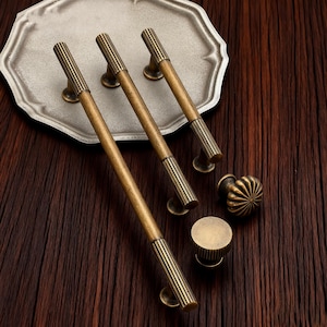 Può includere: Una collezione di ferramenta per mobili in ottone antico, tra cui tre lunghe maniglie a barra, un pomello rotondo e un piccolo pomello cilindrico. La ferramenta presenta un design a coste ed è esposta su un vassoio decorativo argentato e una superficie in legno scuro.