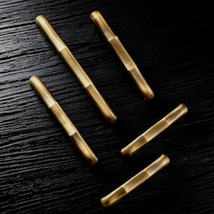 May include: Five brass-coloured, rectangular drawer pulls with a two-tone design. The pulls are arranged diagonally on a dark, textured surface, showcasing their metallic finish. Suitable for cabinet hardware, these pulls are a modern design.