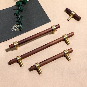 Könnte beinhalten: Eine Auswahl an Holzschrankgriffen mit Messingdetails. Die Griffe haben einen dunkelbraunen, zylindrischen Körper mit Messingringen und T-förmigen Messinggriffen. Die Hardware ist auf einer neutralen beigefarbenen Oberfläche angeordnet.