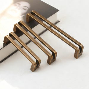 Op de afbeelding: Vier antiek bronzen kastgrepen gerangschikt op een wit oppervlak. Elke greep heeft een rechthoekige handgreep met een dubbel staafontwerp. De grepen zijn in verschillende maten, met een boek op de achtergrond.