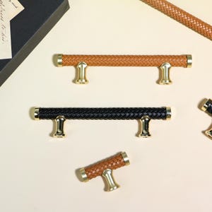 May include: An assortment of cabinet pulls with woven leather-like handles. The pulls are in brown and black, with gold-colored accents. The handles are cylindrical and have a braided texture. The pulls are designed for furniture.