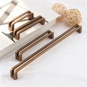 Peut inclure: Plusieurs poignées de meubles de couleur bronze de différentes tailles sont présentées sur une surface blanche. Les poignées ont un design rectangulaire avec une rainure sombre au centre. Deux sphères décoratives tressées sont en arrière-plan.