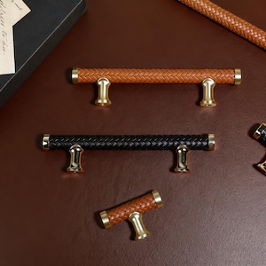 May include: A collection of cabinet pulls with woven leather-like textures in brown and black. The pulls have gold-colored metal accents. The pulls are in various sizes and orientations, suggesting a set for different cabinet doors and drawers.