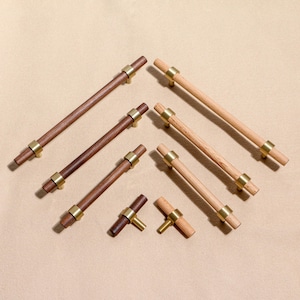 Puede incluir: Una colección de tiradores y pomos de madera para armarios. Los tiradores son rectangulares con un acabado marrón oscuro o claro, acentuados con anillos metálicos de color latón. Los pomos tienen forma de T con un diseño similar. El hardware está dispuesto sobre un fondo neutro.