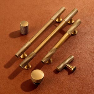 May include: A collection of brass cabinet hardware, including long bar pulls, a T-shaped pull, and two knob styles. The hardware features a ribbed design on the ends and a warm, golden finish. The pieces are arranged on a reddish-brown surface.