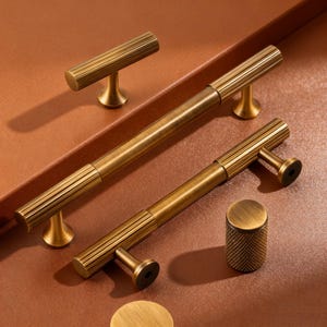 May include: A selection of antique brass cabinet handles and knobs. The hardware has a ribbed design and a warm, aged finish. The pieces are displayed on a brown surface. The cabinet hardware is suitable for furniture.