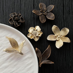 May include: A selection of decorative floral hardware in various styles and finishes. The collection includes flower and leaf-shaped pulls in gold and bronze tones. The hardware is displayed on a dark wooden surface and a white plate.