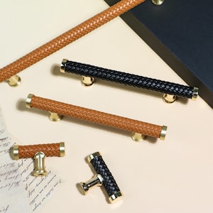Pode incluir: Uma variedade de puxadores e maçanetas de armário com um design de couro trançado. Os puxadores são em tons de marrom e preto, com detalhes dourados. O hardware é projetado para móveis e armários.