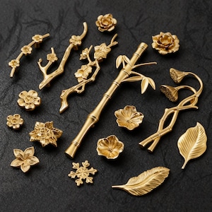 May include: A selection of gold-coloured decorative hardware, including drawer pulls and knobs. The assortment features floral, bamboo, and leaf designs. The hardware is arranged on a dark surface, showcasing the intricate details of each piece.