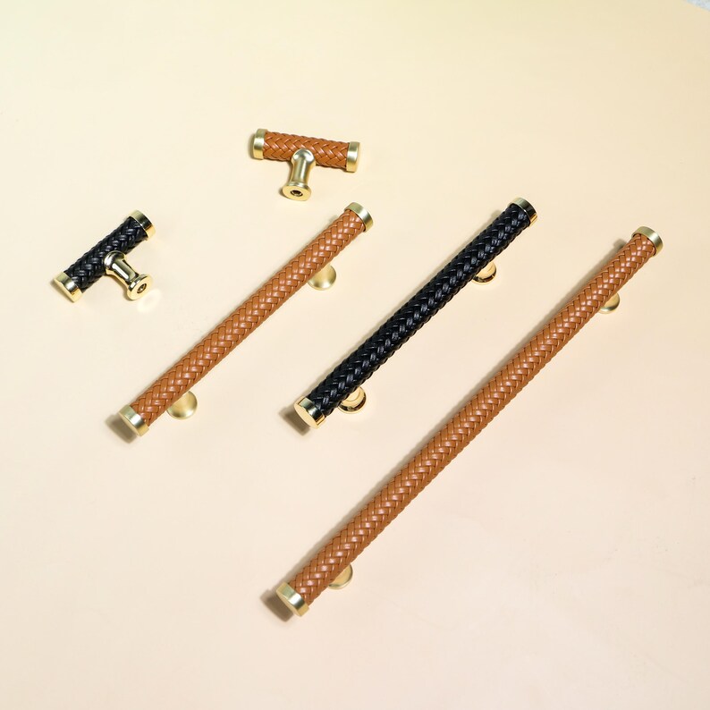 Puede incluir: Una variedad de tiradores y pomos para armarios. El herraje presenta un dise&ntilde;o de cuero trenzado en tonos marrones y negros, con detalles met&aacute;licos dorados. La imagen muestra pomos en forma de T y tiradores largos, dispuestos sobre una superficie beige claro.