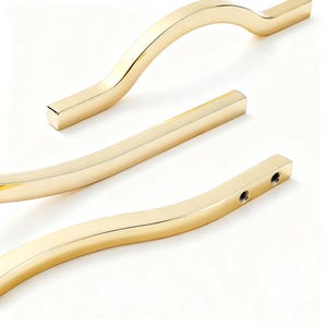 May include: Close-up of three gold-colored cabinet handles. The handles have a curved design, with a rectangular bar in the middle. The handles are made of a shiny, metallic material, and are set against a white background.