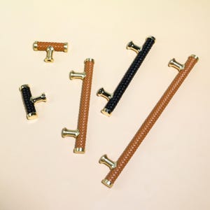 Op de afbeelding: Een assortiment kast- en ladetrekkers in verschillende maten en stijlen. De trekkers hebben geweven leer in zwart en bruin, met goudkleurige hardware. De T-vormige en staafvormige trekkers zijn gerangschikt op een lichtbeige achtergrond.