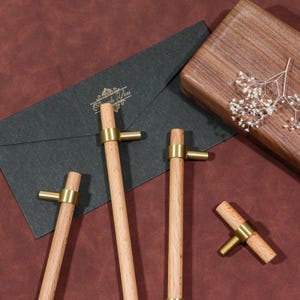 May include: Wooden cabinet pulls with brass handles. The pulls are cylindrical and have a brass handle. A dark grey envelope with gold text and a wooden box are in the background. The background is a dark red-brown colour.