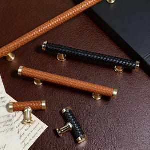 Op de afbeelding: Close-up van verschillende kastgrepen met gevlochten lederachtige handgrepen in bruin en zwart. De grepen hebben goudkleurige accenten en zijn gerangschikt op een bruin oppervlak. Sommige zijn lang, andere korter.