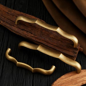 May include: Three antique brass cabinet pulls with curved designs. The pulls are in various shapes, including a wavy and a curved design. The hardware is set against a dark wood background, with a warm, aged finish. These are ideal for adding a vintage touch to furniture.