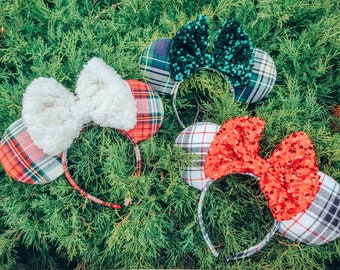May include: Three pairs of festive plaid Minnie Mouse ears with different bows. One pair has a white faux fur bow, another has a red sequin bow, and the third has a green sequin bow.