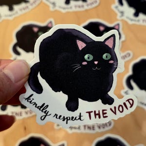 May include: A black cat sticker with green eyes, pink ears, and the phrase "kindly respect THE VOID." The cat is round and cartoonish, with a white border. Other similar stickers are in the background.