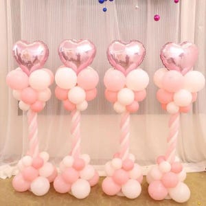 Puede incluir: Cuatro columnas de globos rosas y blancos. Cada columna tiene un globo en forma de corazón en la parte superior, con un grupo de globos rosas y blancos debajo. Las columnas tienen un diseño retorcido rosa y blanco.
