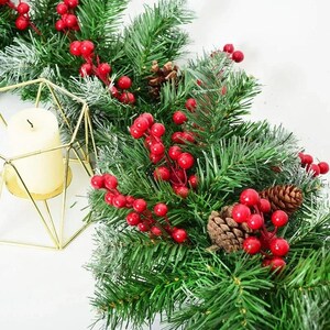 May include: A festive Christmas garland with green pine needles, red berries, and pine cones. A gold geometric candle holder with a cream-colored candle is on the left. The garland is a decorative holiday accent.