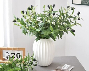 May include: A white, ribbed ceramic vase filled with faux olive branches. The branches have green leaves and black olives. A small wooden calendar shows August 20. A sprig of olives lies on the table.