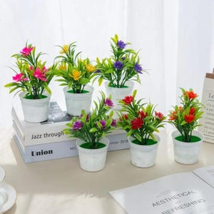 Puede incluir: Una colección de pequeñas plantas artificiales en macetas con flores vibrantes en tonos rosa, amarillo, morado y rojo. Las plantas están en macetas blancas, dispuestas sobre una superficie clara, algunas sobre libros. La escena es brillante y colorida.