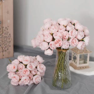 May include: A bouquet of light pink peonies in a clear glass vase with a gold rim. Another smaller bouquet of pink peonies is on the left. The flowers are arranged on a gray surface, with a decorative lantern in the background.