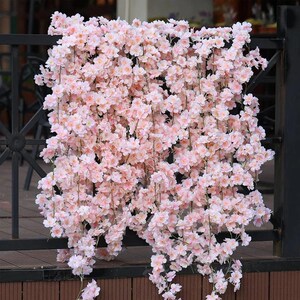 以下が含まれることがあります： 黒い金属の手すりに降り注ぐ、人工のピンク色の桜の花の密集した配置。 花は満開で、柔らかくロマンチックな美学を作り出しています。 花は薄いピンク色で、濃いピンク色のアクセントがいくつかあります。