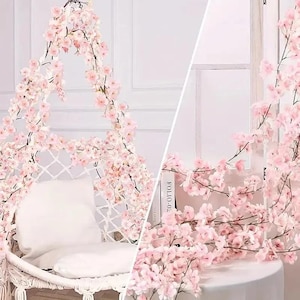 以下が含まれることがあります： ピンクの桜の花のガーランドで飾られた白いマクラメのスイングチェア。白い枕が椅子の中に置かれています。画像の右側には、さらにピンクの花と「FOLIO-02」というテキストの入った本が表示されています。