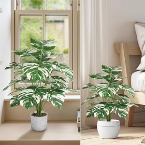 May include: Two artificial plants with large green and white variegated leaves. One plant is in a speckled grey pot, the other in a white pot. Both are placed near a window and a bed.