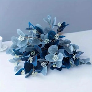 May include: A bouquet of artificial eucalyptus leaves in shades of blue and gray. The leaves have a velvety texture and are accented with small white flower clusters. The arrangement is set against a light blue background, creating a calming aesthetic.