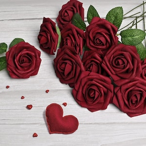May include: A display of deep red artificial roses with green leaves, arranged on a light wood-grain surface. A felt heart and small heart-shaped accents are also present, creating a romantic theme. The roses are clustered together, suggesting a bouquet or decorative arrangement.
