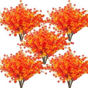 Puede incluir: Cinco ramos de hojas de eucalipto artificiales naranjas y amarillas. El follaje artificial tiene una mezcla de hojas naranjas y amarillas con tallos marrones oscuros. Las plantas artificiales están dispuestas en un patrón simétrico sobre un fondo blanco.