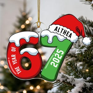 May include: Christmas ornament featuring the numbers 67 in red and green, with a Santa hat. The ornament reads "THE YEAR OF" and "2025" with the name "ALTHEA" on the hat. The ornament hangs from a gold string.