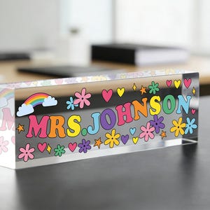Puede incluir: Un bloque acrílico transparente con el nombre "MRS. JOHNSON" en letras coloridas de estilo retro. El diseño incluye un arcoíris, flores, corazones y estrellas. El bloque rectangular está sobre una superficie oscura.