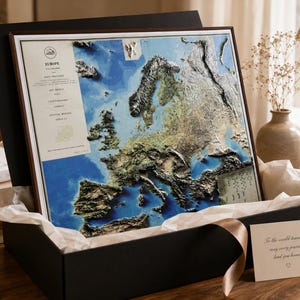 Op de afbeelding: Een gedetailleerde 3D-kaart van Europa, ingelijst en gepresenteerd in een zwarte geschenkdoos met een lint. De kaart toont blauwe, groene en bruine topografische details. Een klein kaartje leest: "To the world traveler, may every journey lead you home."