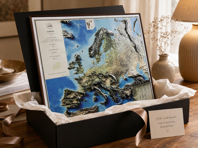 Op de afbeelding: Een gedetailleerde 3D-kaart van Europa, ingelijst en gepresenteerd in een zwarte geschenkdoos met een lint. De kaart toont blauwe, groene en bruine topografische details. Een klein kaartje leest: "To the world traveler, may every journey lead you home."