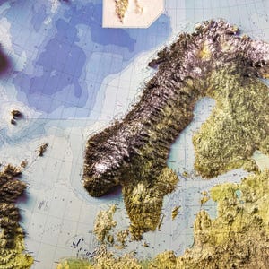 Op de afbeelding: Een gedetailleerde reli&euml;fkaart van Noord-Europa, met de Scandinavische schiereiland en de omliggende gebieden. De kaart toont getextureerde, verhoogde landmassa's in bruin- en goudtinten, tegen een blauw-witte oceaanachtergrond. Rasterlijnen en labels zijn zichtbaar.