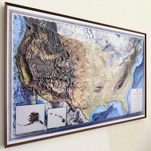 May include: A framed wall map of the United States featuring a three-dimensional relief design. The map displays topographical mountain ranges in dark brown and tan, with blue ocean depths. Insets show Alaska and Hawaii in the lower left corner.