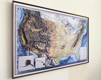 May include: A framed 3D relief map of the United States, showcasing topographical details with varying shades of brown, green, and blue. The map includes insets of Alaska and Hawaii. The frame is a dark brown color.