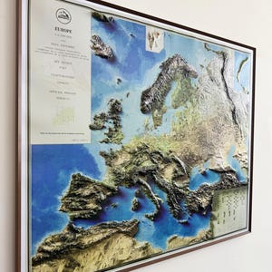 May include: A framed 3D relief map of Europe, showcasing the continent's topography with detailed elevations. The map features shades of blue for water and varying earth tones for land, with text in the upper left corner. The frame is brown.