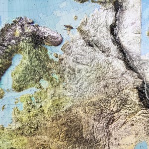 Op de afbeelding: Een gedetailleerde reli&euml;fkaart van Europa, die de gevarieerde topografie van het continent laat zien. De kaart toont tinten groen, bruin en wit, die bergen, vlaktes en wateren vertegenwoordigen. De kaart heeft een blauwe achtergrond met rasterlijnen.