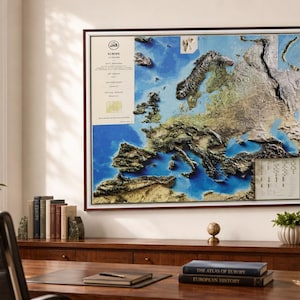Op de afbeelding: Een ingelijste reli&euml;fkaart van Europa, die de topografie van het continent weergeeft met gedetailleerde texturen en kleuren. De kaart staat tegen een blauw-witte achtergrond, met een donkerbruine lijst. De kaart wordt weergegeven op een houten bureau met boeken en een lamp.