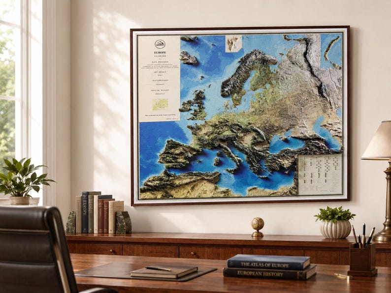 Op de afbeelding: Een ingelijste reli&euml;fkaart van Europa, die de topografie van het continent weergeeft met gedetailleerde texturen en kleuren. De kaart staat tegen een blauw-witte achtergrond, met een donkerbruine lijst. De kaart wordt weergegeven op een houten bureau met boeken en een lamp.